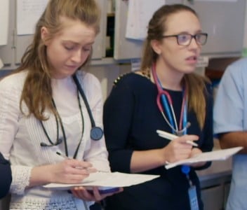 Two female doctors with stethoscopes take notes using pens and pads